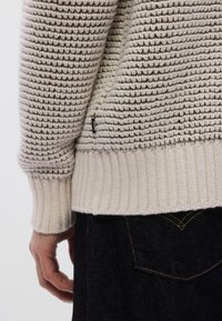 Ανοιχτό μπεζ πλεκτό πουλόβερ με υφασμένο μοτίβο, ριμπ μανσέτες και τελείωμα. Διαθέτει μια μικρή μαύρη ετικέτα στο πλάι. Φοριέται με σκουρόχρωμο τζιν.