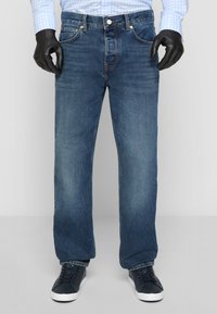Blue denim jeans with a straight cut, five pockets, and metal button closure. Worn with black sneakers and a checked blue shirt.