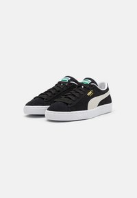 Baskets en suède noir avec semelle en caoutchouc blanche, présentant une bande latérale crème et des accents de logo verts. Lacets plats et design bas.
