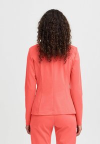 Blazer cintré de couleur corail avec manches longues, tissu lisse, design structuré et coutures au dos pour une définition de la silhouette, assorti à un pantalon coordonné.
