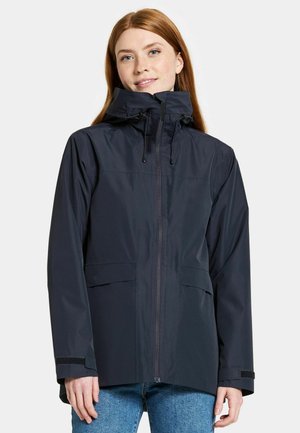 Vrouw draagt een marineblauwe waterdichte jas met een rits en hoge kraag en verstelbare capuchon, gecombineerd met blauwe jeans.