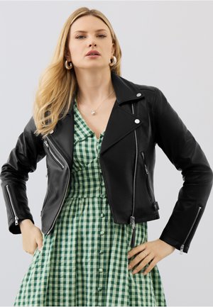 Blonde Frau mit silbernen Creolen, einer Herz-Halskette, einer schwarzen Lederjacke und einem grünen karierten, geknöpften Kleid.