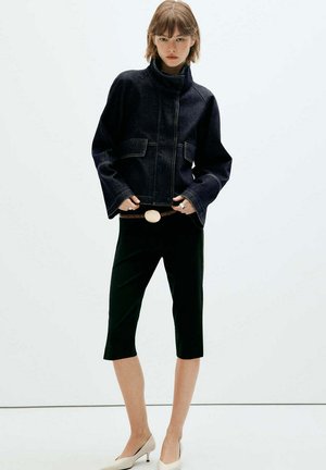 Mujer con chaqueta vaquera oscura, pantalones cortos negros con cinturón blanco y zapatos beige de tacón bajo, de pie frente a un fondo liso.