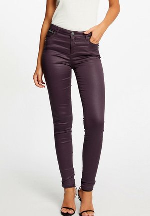 Vrouw draagt een donkerpaarse skinny broek met hoge taille en ritssluitingen in de zakken, een wit topje en zwarte open-toe hakken, staand voor een witte achtergrond.
