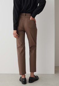 Bruine enkel-lange broek met een rechte snit, voorzien van twee achterzakken en een gladde textuur. Gecombineerd met zwarte instappers en een zwart top.