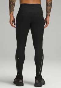 Fekete sportleggingset viselő férfi, fényvisszaverő csíkokkal és fekete futócipőben, hátulról látható, szürke háttér előtt.