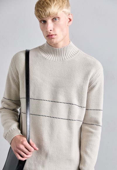 Beige stickad tröja med hög ribbad krage och svarta horisontella ränder. Modellen håller en svart axelväska. Slät textur.