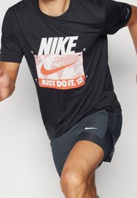 Černé sportovní tričko s velkým bílým nápisem "NIKE" a designem "JUST DO IT" v oranžové a bílé barvě s grafickými prvky.