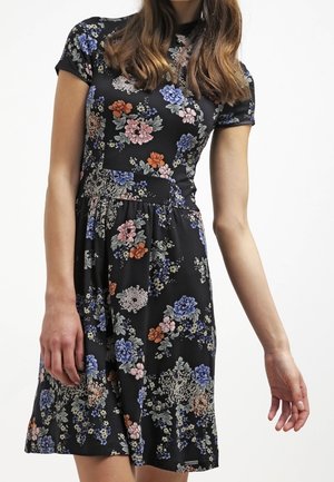 Kobieta ubrana w czarną sukienkę z krótkim rękawem, wysokim kołnierzem i kolorowymi kwiatowymi wzorami w odcieniach niebieskiego, różowego, pomarańczowego i białego.