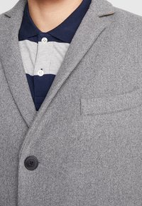 Blazer grigio con un misto di lana strutturato, con un singolo bottone nero e una tasca sul petto. Sotto, è visibile una polo a righe blu navy e grigie.