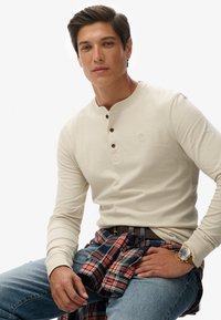 Camisa Henley de manga larga en color beige con tres botones oscuros, combinada con una camisa de franela de cuadros anudada en la cintura. Usada con jeans azules y un reloj.