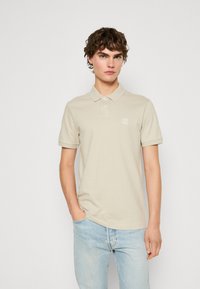 Μπεζ πόλο μπλούζα φτιαγμέ�νη από απαλό βαμβάκι, με κλασικό γιακά, κοντά μανίκια και μια μικρή ετικέτα λογότυπου στο στήθος. Συνδυασμένη με ανοιχτό μπλε τζιν.