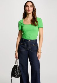 Groene geribbelde top met korte mouwen en een vierkante halslijn, gecombineerd met donkerblauwe wijde jeans en een gestructureerde zwarte handtas.