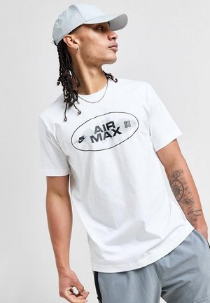Persona che indossa una t-shirt bianca Nike Air Max e un cappellino grigio chiaro, guarda di lato mostrando un tatuaggio sull'avambraccio sinistro, su sfondo uniforme.