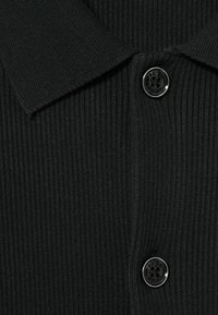 Cămașă polo neagră cu rigle, dotată cu guler plat și închidere cu nasture, având un nasture rotund, negru, pe care este afișat un logo.
