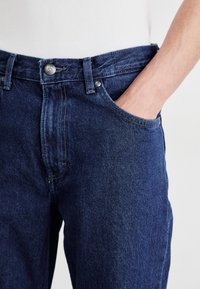 Isik, kellel on seljas tume sinine teksade paar, käsi eesolevas taskus, vöökohast ja osaliselt nähtav valgest topist.