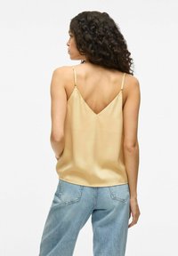 Camiseta de satén en un tono beige claro con diseño de espalda en V y tirantes delgados ajustables, combinada con unos vaqueros de mezclilla azul claro. Textura suave.