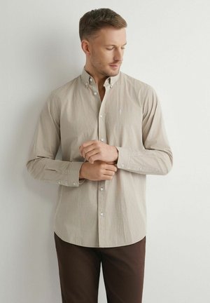 Mężczyzna poprawiający mankiet beżowej koszuli w kratę zapinanej na guziki, ubrany w ciemnobrązowe spodnie, stoi na tle jasnej, gładkiej ściany.