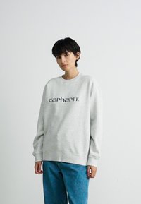 Persoon met kort, donker haar, gekleed in een lichtgrijze Carhartt sweatshirt en blauwe jeans, staande tegen een effen witte achtergrond.