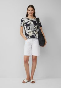 Shorts blancs et un haut à imprimé floral noir avec des feuilles beiges. Accessoirisé avec un sac en crochet noir et des sandales blanches.