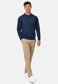 Tamno plavi dugih rukava polo pulover s kragnom na gumbe, uparen s bež chinos hlačama i bijelim trenerkama. Glatka tkanina, fitted dizajn.