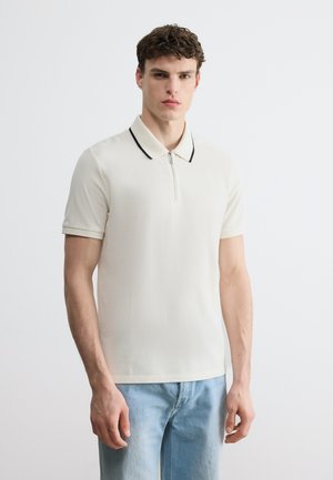 Jonge man die een crème poloshirt met korte mouwen draagt, met zwarte rand op de kraag, en lichtblauwe spijkerbroek, staand tegen een effen achtergrond.