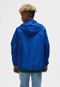 Blå hættejakke med elastiske manchetter, letvægtstof og en glat tekstur. Båret over en grøn skjorte med blå denimjeans.