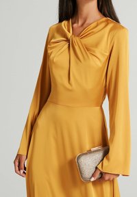 Gul långärmad klänning med en vriden knut vid halsringningen, slät yta och en figursydd silhuett. Silverfärgad metallclutch hålls i handen.
