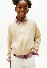 Beige gestrickter Pullover mit einem weißen Herzdesign, das den Text "TOMMY HILFIGER" trägt. Dazu eine rosa karierte Bluse und cremefarbene Hose.