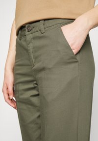 Khaki kalhoty z lehkého materiálu s bočními kapsami, zapínáním na knoflík a čistým, upraveným střihem. Žádné viditelné vzory nebo akcenty.