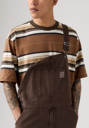 Homme portant un t-shirt rayé marron et une salopette marron avec une poche avant, montrant ses avant-bras tatoués devant un fond uni.