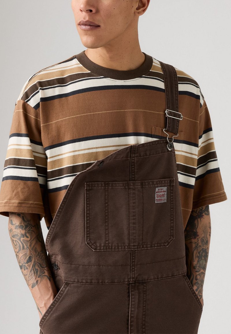 Man in bruin gestreept T-shirt en bruine overall met een voorzak, met getatoeëerde onderarmen tegen een effen achtergrond.