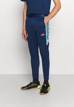 Personne portant un pantalon de survêtement Adidas bleu marine avec des bandes latérales bleu clair et des chaussures de football en salle Adidas Copa noires avec des détails jaune fluo.