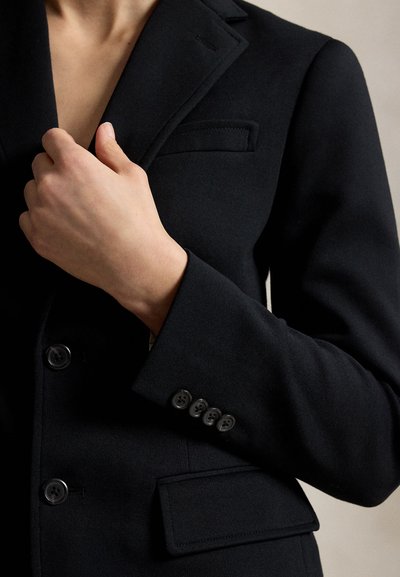 Persoon die de revers van een zwarte op maat gemaakte blazer met knoopmanchetten en een klepzak aan de voorkant aanpast.