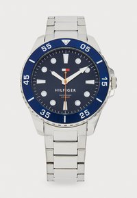 Montre en acier inoxydable argenté avec une lunette bleue, un cadran marine et des marqueurs lumineux. Dispose d'une aiguille des secondes orange et d'un bracelet à maillons.