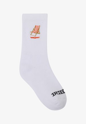Weißer Socken mit einem bestickten orange-beigen gestreiften Liegestuhl und Text darunter sowie schwarzem Schriftzug "IRIEDAILY" nahe der Fußspitze.