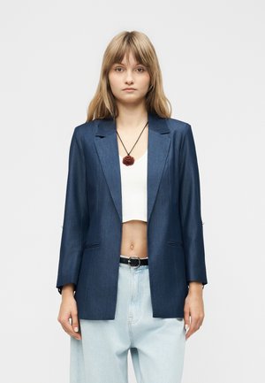 Mladá žena stojaca, oblečená v tmavomodrom blejzeri, bielom krátkom topu, svetlomodrých džínsoch s čiernym opaskom a červeno-ružovým náhrdelníkom.