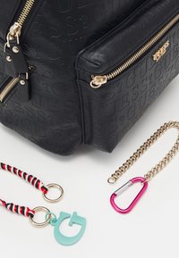 Černá taška s texturou, zlatými zipy a logem. Obsahuje červeně a černě pruhované popruhy, tyrkysovou visačku a růžový karabiner.