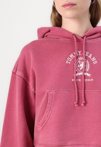 Felpa in cotone rosa con tasca a marsupio, cappuccio con coulisse e logo bianco che presenta un leone e il testo: "TOMMY JEANS EST. 1985."