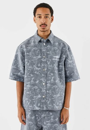 Man die een grijs overhemd met korte mouwen en paisley-print draagt, gecombineerd met een bijpassende broek en een wit geborduurd logo op de borst.