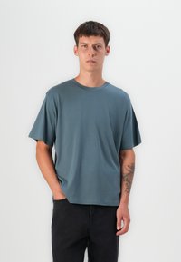 Tricou cu mânecă scurtă, de tip crew-neck, într-o nuanță de teal estompat, realizat dintr-un material moale și neted. Are o croială lejeră și o ușoară întindere, asortat cu pantaloni întunecați.