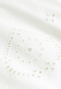 Fehér anyag finom kivágott hímzett virág- és geometriai mintákkal, amelyek texturált és légies mintázatot hoznak létre.