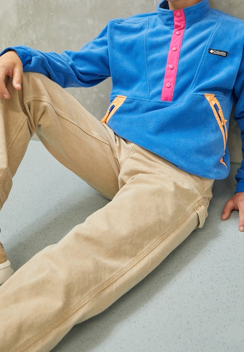 Pullover in pile blu con patta a bottoni rosa e tasche con zip arancioni, abbinato a pantaloni beige. Tessuto morbido e vestibilità rilassata.