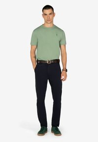 T-shirt verde a maniche corte, pantaloni blu navy, cintura marrone, scarpe verdi. Presenta un piccolo logo sulla maglietta e una cintura strutturata.