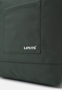 Zielona torba materiałowa o teksturowanej powierzchni, z wyraźną prostokątną kieszenią i białym haftem logo "Levi's" na dolnej krawędzi.
