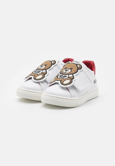 Hvite barnesko med rødt interiør, med store bamse-patcher merket "leketøy" på de justerbare frontstroppene.