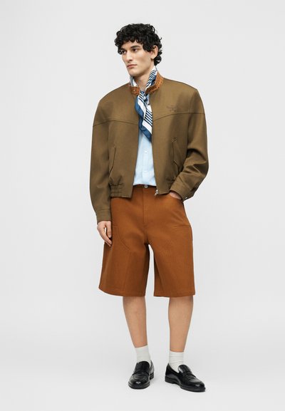 Άνδρας που στέκεται φορώντας καφέ μπουφάν bomber, ανοιχτό μπλε πουκάμισο, καφέ σορτς μέχρι τα γόνατα, μπλε φουλάρι με σχέδια στον λαιμό, λευκές κάλτσες και μαύρα παπούτσια.