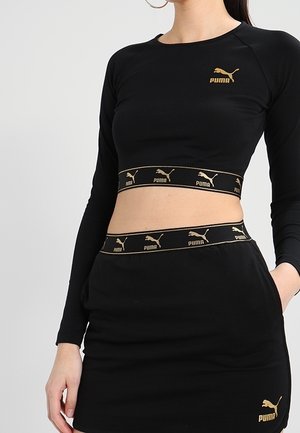 Kobieta ubrana w czarny crop top i spódnicę marki Puma z złotymi logo Puma oraz elastycznymi pasami z logo na obu elementach garderoby.