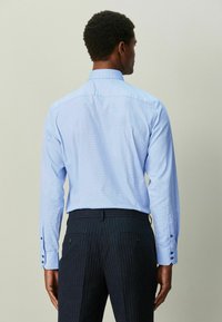 Lyseblå mønstret skjorte med knapper, spredt krave, lange ærmer og mørke, stribede bukser, set bagfra.