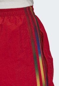 Červené šortky vyrobené z lehkého materiálu se svislými vícestupňovými proužky po stranách a kapsou na zip. Texturovaný povrch.